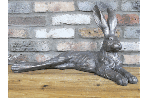 Metallic laying rabbit sculpture with elongated body and upright ears, placed on a wooden surface against a brick wall.