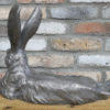 Metallic laying rabbit sculpture with detailed fur and long ears, placed on a wooden surface against a brick wall background.