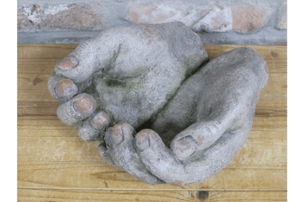 Weathered stone hands sculpture with detailed fingers, placed on a wooden surface against a brick wall background.