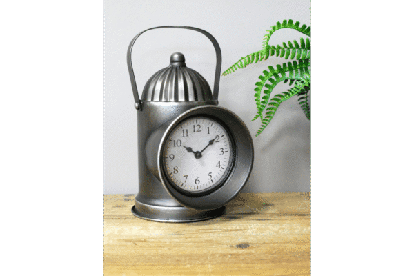 Vintage metal lantern clock with a round face and black numerals, placed on a wooden surface with a green plant in the background.