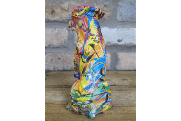 Colourful graffiti-style dog sculpture with vibrant paint splashes and abstract designs, placed on a wooden surface against a brick wall background.