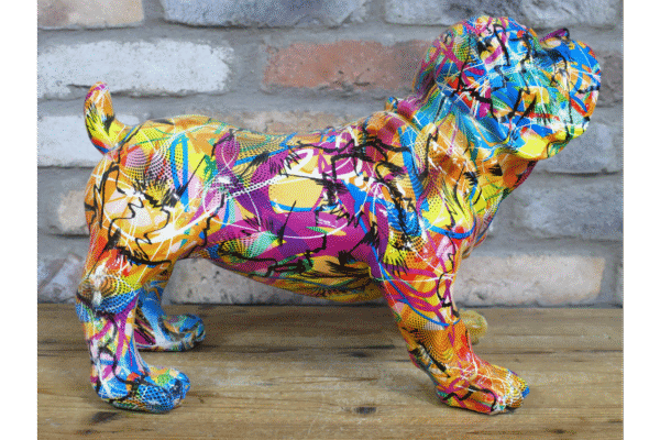 Colourful graffiti-style dog sculpture with vibrant paint splashes on a wooden surface against a brick wall.