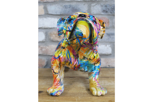 Colourful graffiti-style dog sculpture with vibrant paint splashes and abstract patterns, sitting on a wooden surface against a brick wall background.