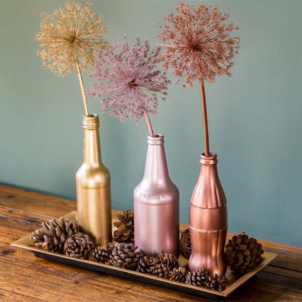 Decorative bottles painted in gold, pink, and copper with dandelion seed heads, surrounded by pinecones on a wooden tray.