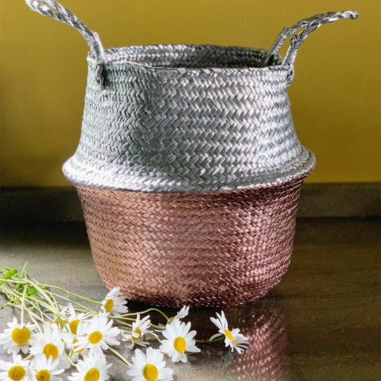 Decorative metallic basket with woven texture and two handles, placed on a wooden surface with daisies in front.