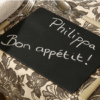 Chalkboard with 'Philippa Bon appétit!' written in white chalk on a decorative tablecloth.