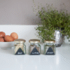 Glass jars labeled Bay, Cumin, and Chives with eggs and a potted herb in the background.