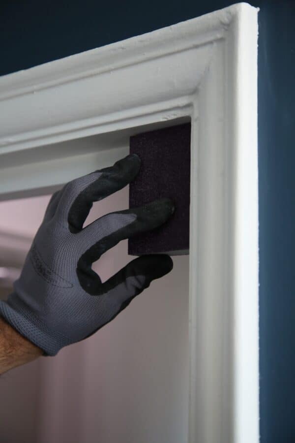 Close-up of a hand wearing a grey glove applying a black sanding sponge to a white door frame for smooth finishing.