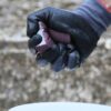 Close-up of a gloved hand holding a purple sanding block outdoors, with water droplets on the hand and background of gravel.