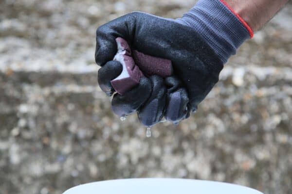 Close-up of a gloved hand holding a purple sanding block outdoors, with water droplets on the hand and background of gravel.