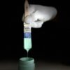 Close-up of a hand in a white glove holding a blue and green paintbrush above a small open container of mint green paint.