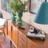 Stylish wooden sideboard decorated with green glass vases and a modern blue lamp, set in a bright room with large windows.