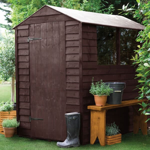 Wooden garden shed painted in dark brown with a sloped roof, situated in a lush garden setting with potted plants and a wooden bench nearby.