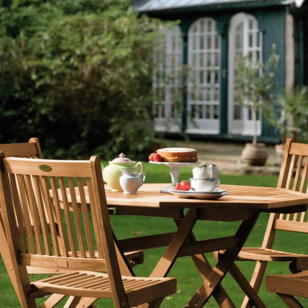 Outdoor garden scene with wooden table and chairs set for tea, surrounded by greenery and a blue garden shed in the background.