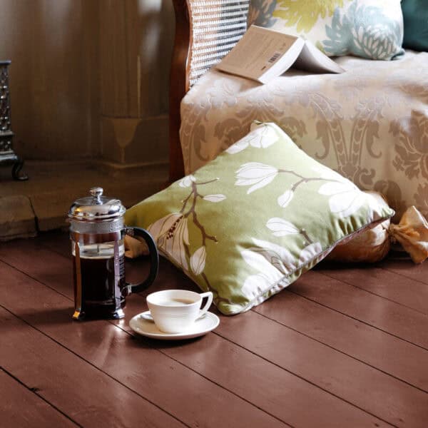 Decorative pillow with leaf pattern on wooden floor next to a coffee pot and cup in a warm, inviting living space.