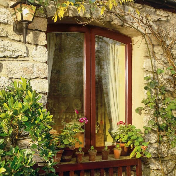 A charming window with a wooden frame set in a stone wall, surrounded by lush green plants and flowering pots, creating a cozy outdoor scene.