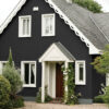 Stylish black house with white window frames and a white porch, surrounded by lush greenery and flowering plants, showcasing exterior wall decoration and masonry paint.