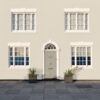 Front view of a white house with four symmetrical windows and a central door, decorated with potted plants on a paved walkway.