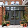 A contemporary conservatory with grey window frames and a glass roof, attached to a brick house, surrounded by potted plants and outdoor paving.