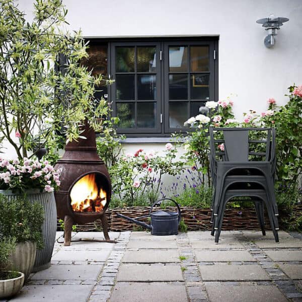 Cozy outdoor garden patio featuring a fireplace, stacked chairs, and lush flowering plants in a residential setting.