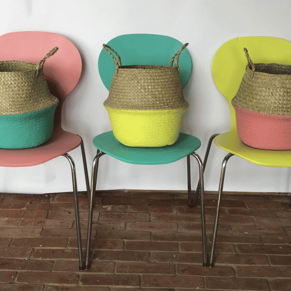 Three woven storage baskets with handles, each painted in different pastel colours, placed on vibrant chairs in a modern interior setting.