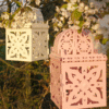 Decorative lanterns hanging from blossoming tree branches, featuring intricate cut-out patterns in pastel pink and cream colors.